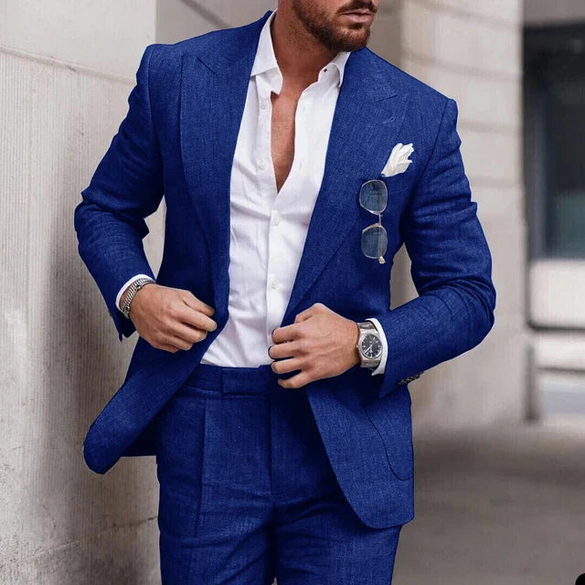 Homme en costume bleu élégant, chemise blanche ouverte, lunettes de soleil accrochées, montre-bracelet argentée, style mode masculine chic.