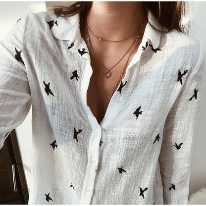 Chemise blanche en lin avec motifs d'oiseaux noirs, col ouvert, portée par une femme. Style décontracté, tendance mode féminine, accessoires minimalistes.