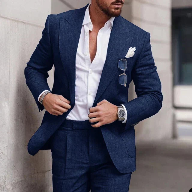 Homme en costume bleu élégant, chemise blanche ouverte, lunettes de soleil accrochées, montre-bracelet argentée, style mode masculine moderne.