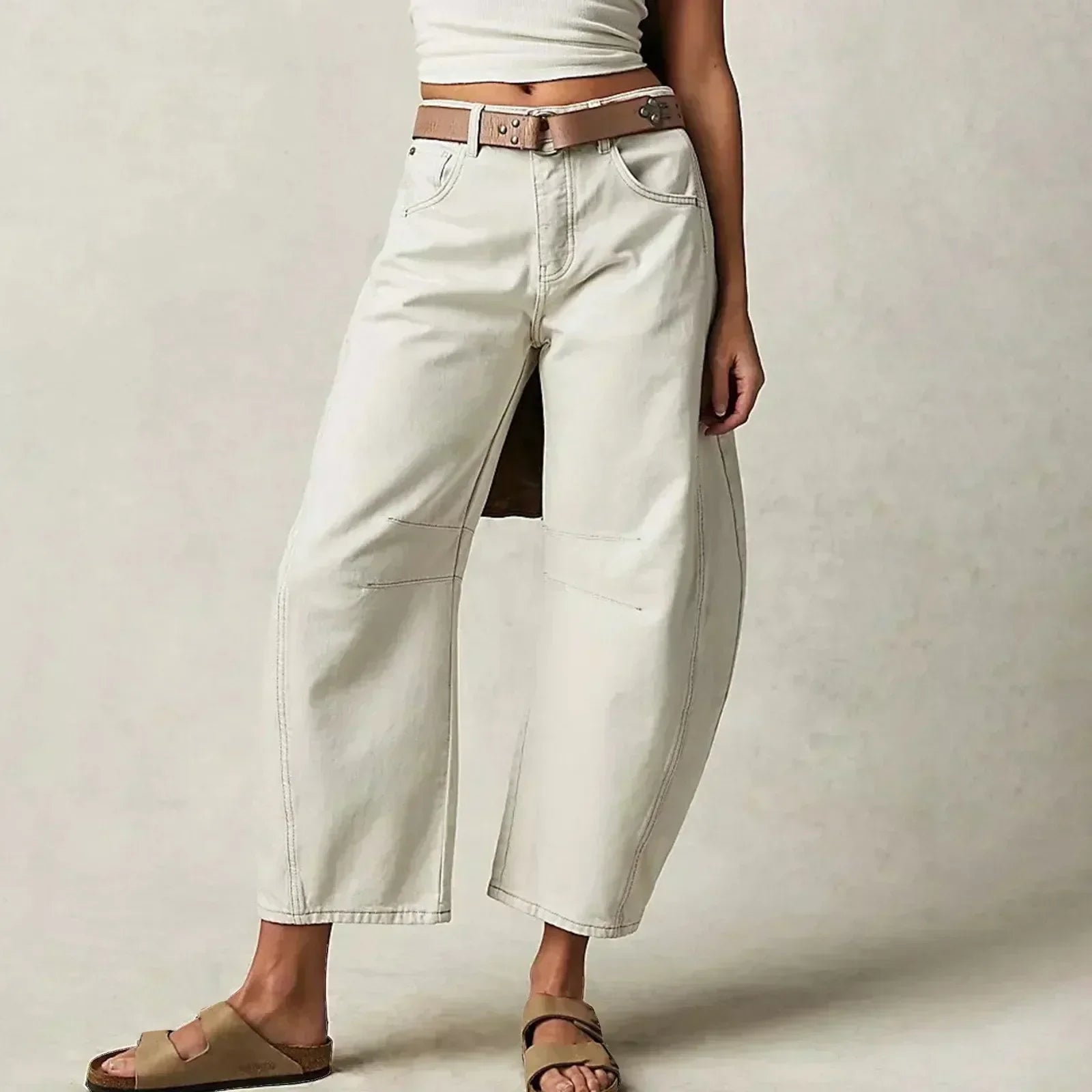 Jeune femme portant un jean blanc ample avec ceinture marron et sandales, posant contre un fond neutre. Mode décontractée, tendance minimaliste.