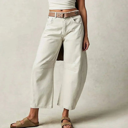 Jeune femme portant un jean blanc ample avec ceinture marron et sandales, posant contre un fond neutre. Mode décontractée, tendance minimaliste.