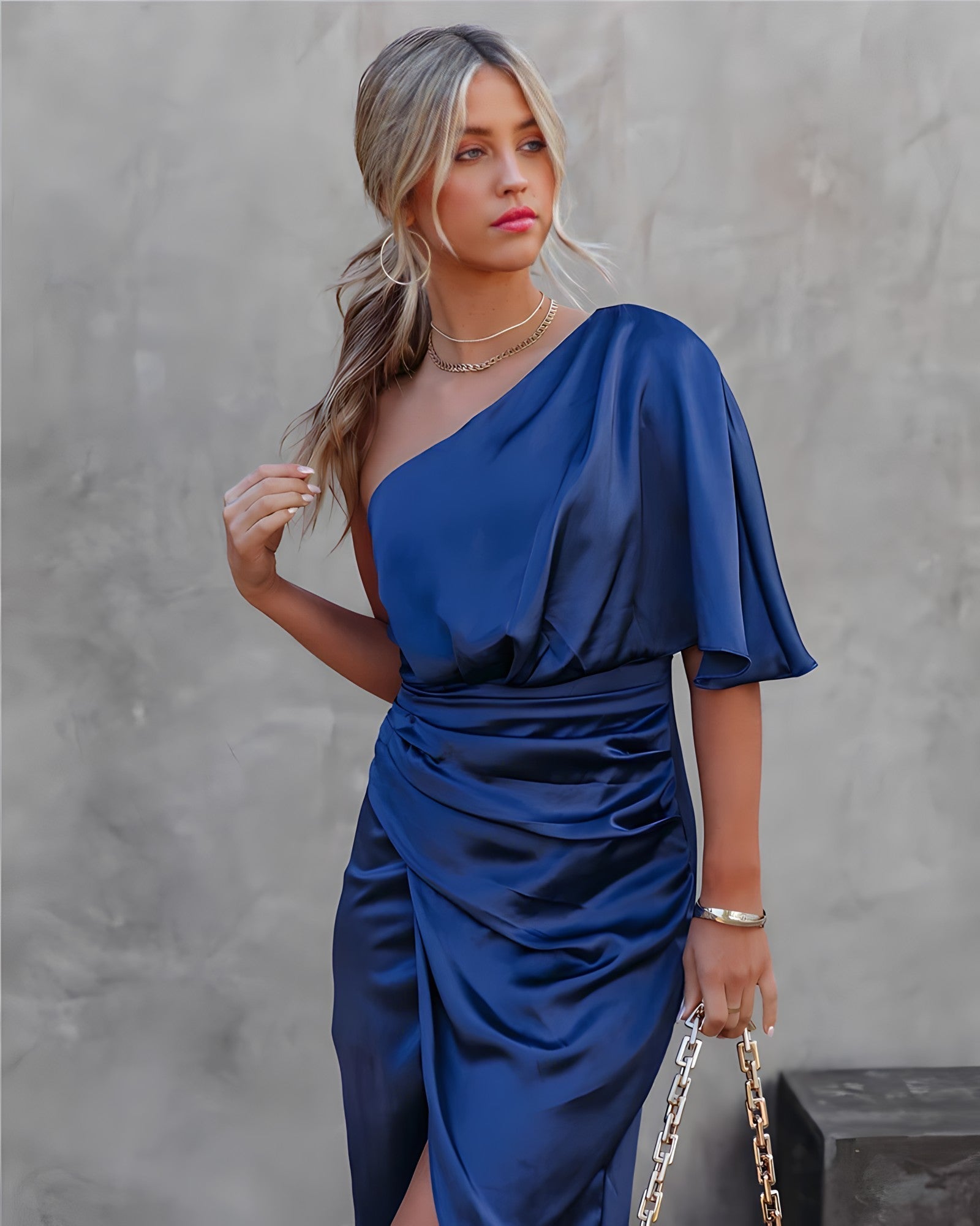 Femme en robe bleue satinée asymétrique, tenant un sac à chaîne dorée, posant devant un mur gris. Mode élégante, style chic, tenue soirée.