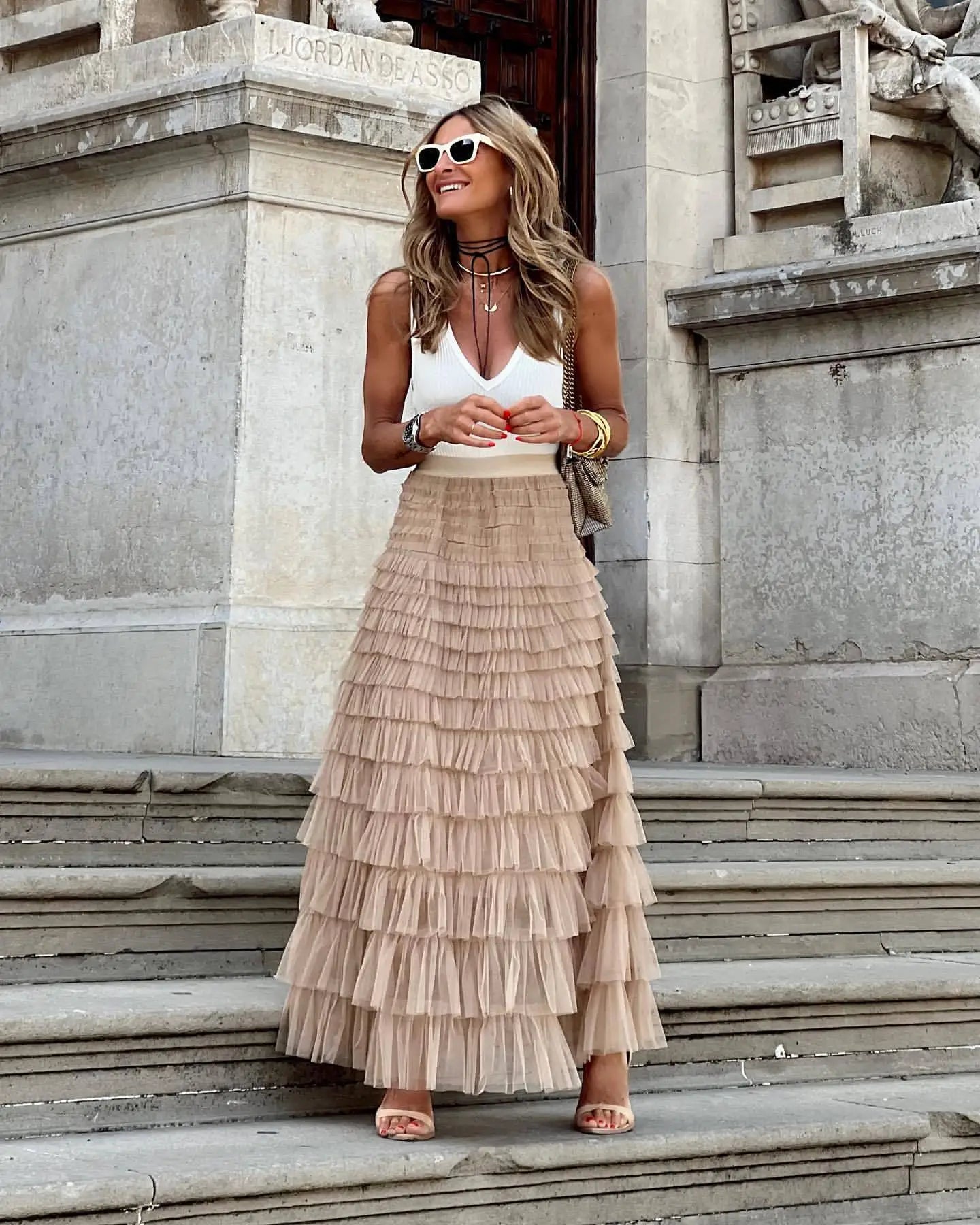 Femme souriante en jupe longue à volants beige et débardeur blanc, posant sur des marches en pierre, lunettes de soleil blanches, style chic urbain.