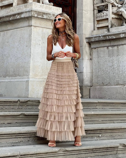 Femme souriante en jupe longue à volants beige et débardeur blanc, posant sur des marches en pierre, lunettes de soleil blanches, style chic urbain.