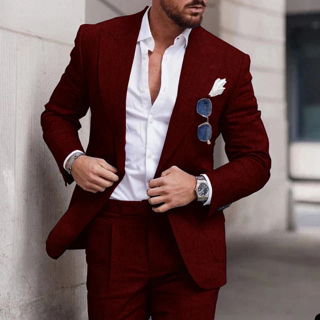 Homme en costume rouge élégant, chemise blanche ouverte, lunettes de soleil accrochées, montre-bracelet argentée, style mode masculine moderne.