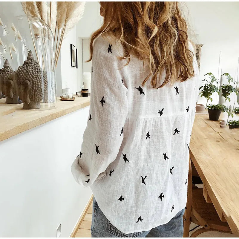 Femme portant une chemise blanche à motifs d'oiseaux noirs, debout dans un intérieur moderne avec des plantes et des sculptures bouddhistes.
