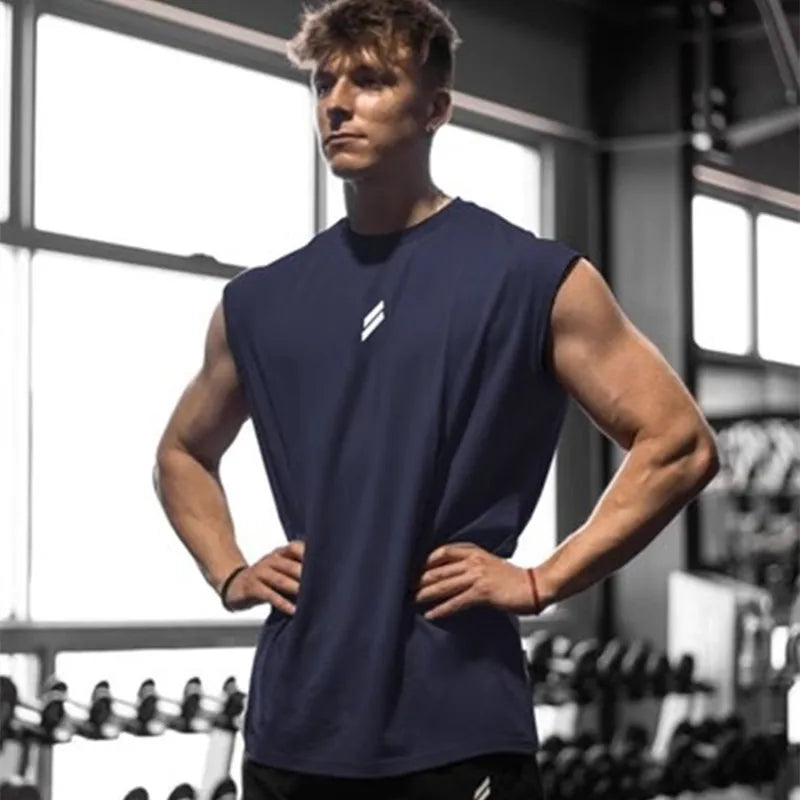 Ung mand i fitnesscenter iført mørkeblå tanktop, står med hænderne på hofterne foran vægtstativ. Styrketræning, fitness, muskelopbygning.