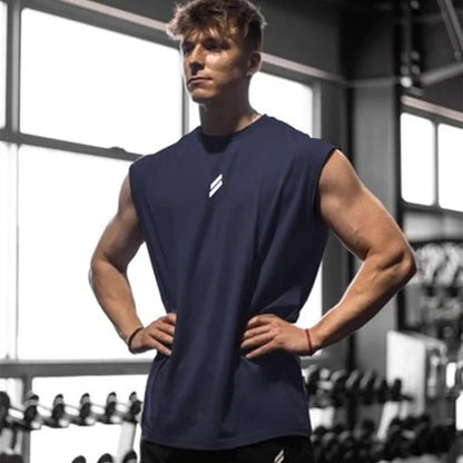 Ung mand i fitnesscenter iført mørkeblå tanktop, står med hænderne på hofterne foran vægtstativ. Styrketræning, fitness, muskelopbygning.
