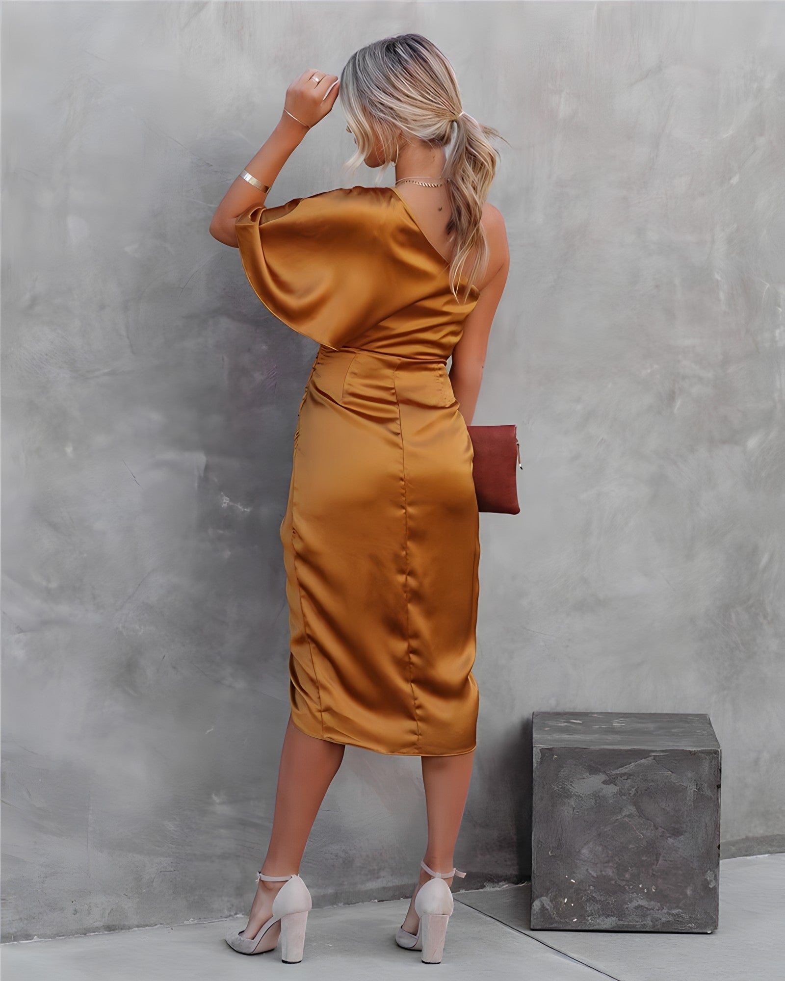 Femme en robe satinée dorée, dos tourné, tenant une pochette marron, posant contre un mur gris. Mode élégante, tenue de soirée chic.