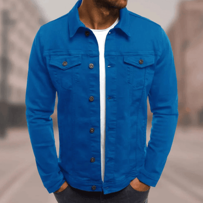 Homme portant une veste en jean bleu vif, chemise blanche en dessous, debout dans une rue floue. Mode masculine, style décontracté.