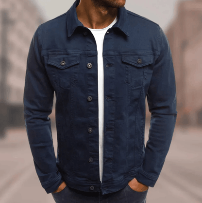 Homme portant une veste en jean bleu foncé, chemise blanche en dessous, debout dans une rue floue. Mode masculine, style décontracté, tendance.