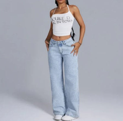 Femme portant un débardeur blanc avec texte, jean large bleu clair, posant debout. Mode décontractée, tendance streetwear, style moderne.
