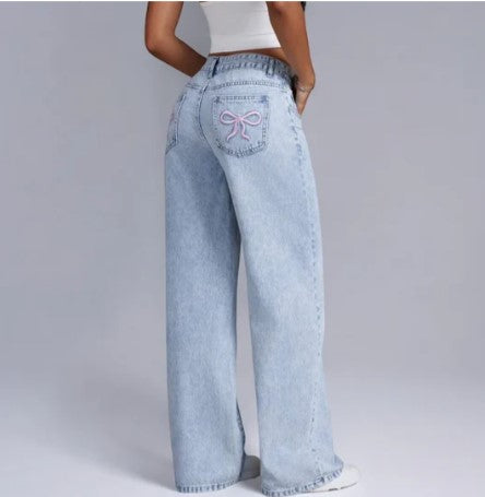 Jeans larges en denim bleu clair avec motif nœud rose sur la poche arrière, portés par une femme de dos, style décontracté et tendance.