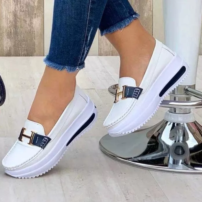 Chaussures mocassins blanches pour femmes avec semelles compensées, ornées d'un détail métallique doré en forme de H, portées avec un jean bleu.
