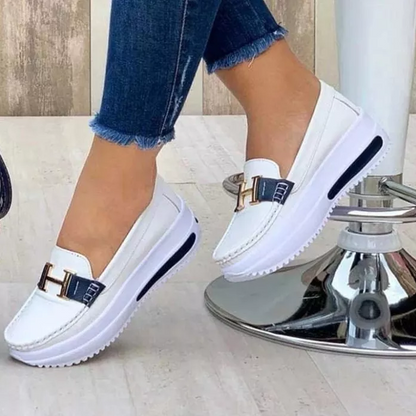 Chaussures mocassins blanches pour femmes avec semelles compensées, ornées d'un détail métallique doré en forme de H, portées avec un jean bleu.