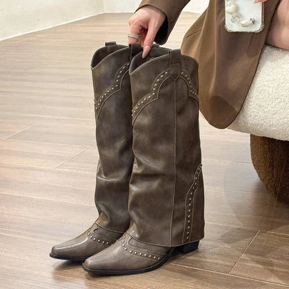 Brune cowboystøvler med nitter på trægulv, holdt af person i brun jakke. Trendy fodtøj, westernstil, mode til kvinder.