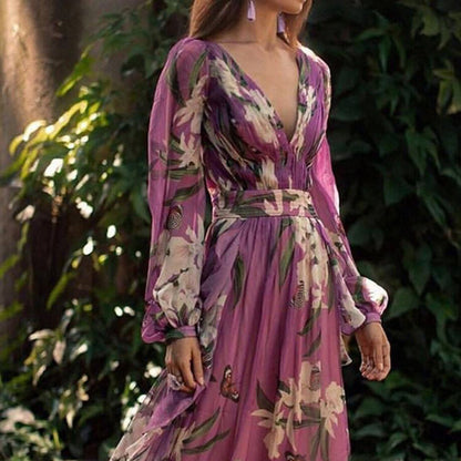 Robe longue violette à motifs floraux, manches longues bouffantes, portée par une femme dans un jardin verdoyant. Mode élégante et tendance.