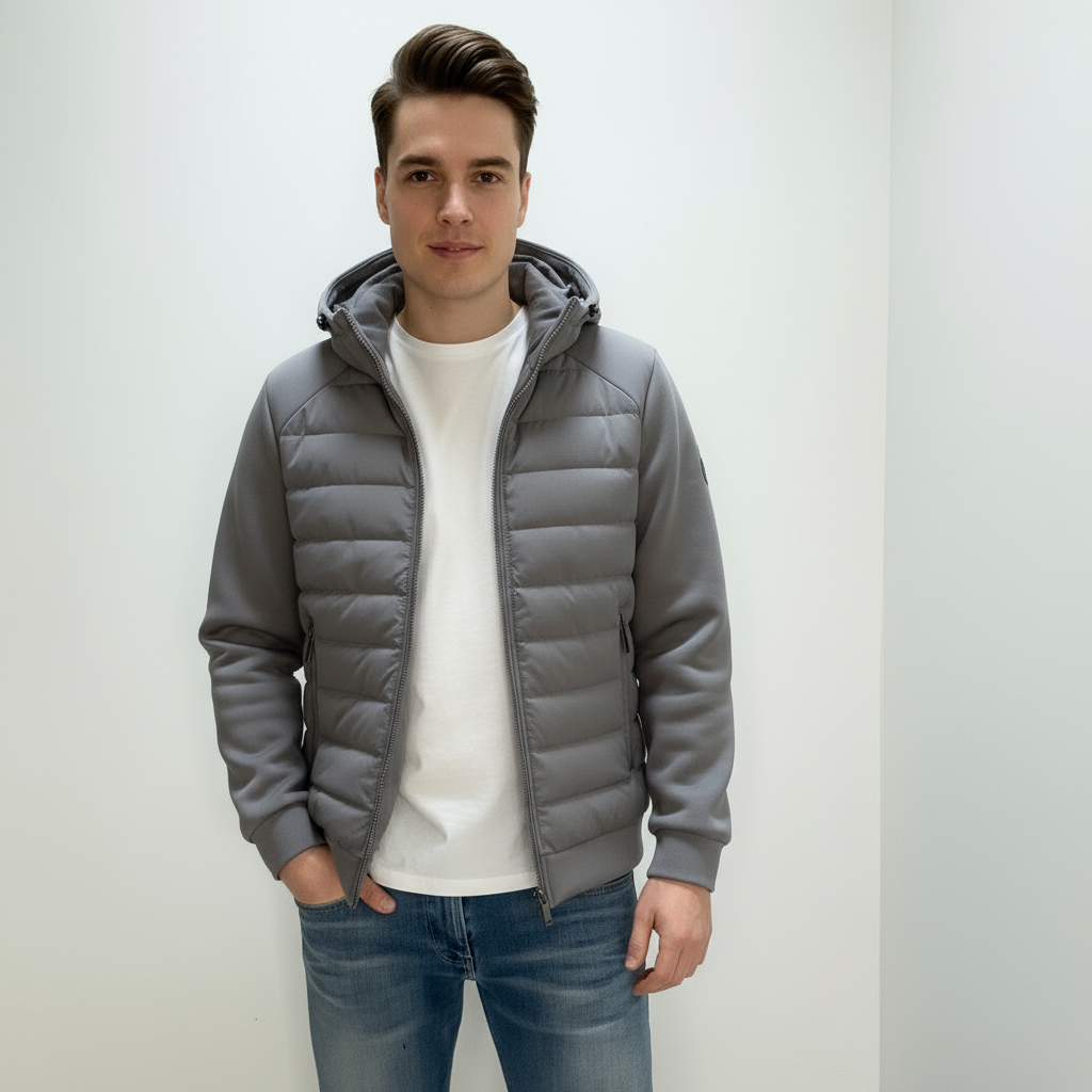Ung mand iført grå hættejakke og blå jeans, står mod hvid baggrund. Moderne herremode, afslappet stil, vinterjakke, casual outfit.