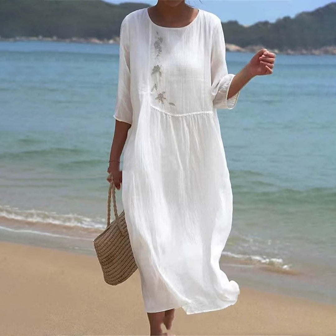 Robe blanche d'été en lin, manches trois-quarts, broderie florale, portée sur une plage ensoleillée avec sac en osier, mer et collines en arrière-plan.