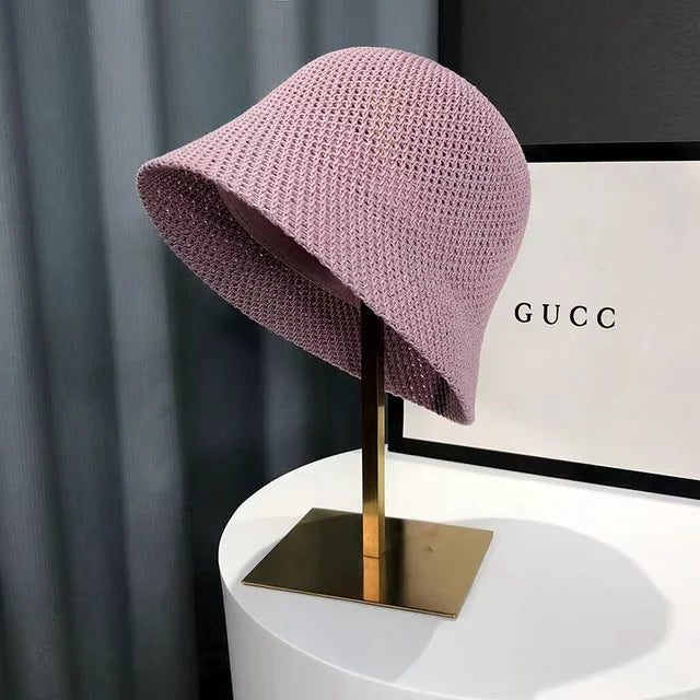 Chapeau cloche en tricot rose sur support doré, devant une boîte Gucci. Accessoire de mode élégant et tendance, idéal pour l'été.