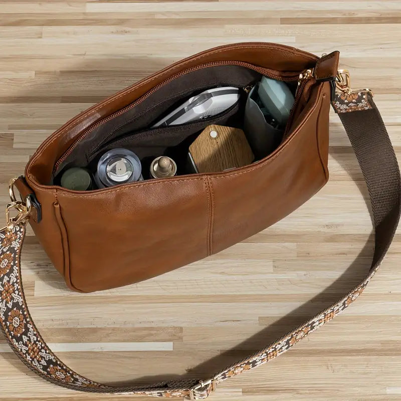 Sac à bandoulière en cuir marron ouvert, montrant des articles de toilette et accessoires à l'intérieur, posé sur une table en bois clair.