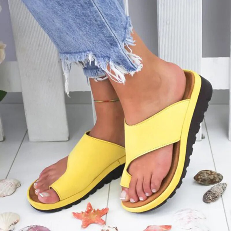 Gule damesandaler med sorte såler, båret af en person i afklippede jeans. Sandalerne er omgivet af muslingeskaller og en søstjerne på hvidt gulv.
