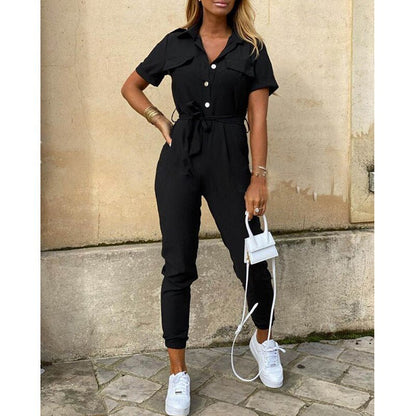 Femme portant une combinaison noire élégante avec ceinture, baskets blanches et petit sac à main blanc, posant devant un mur beige.