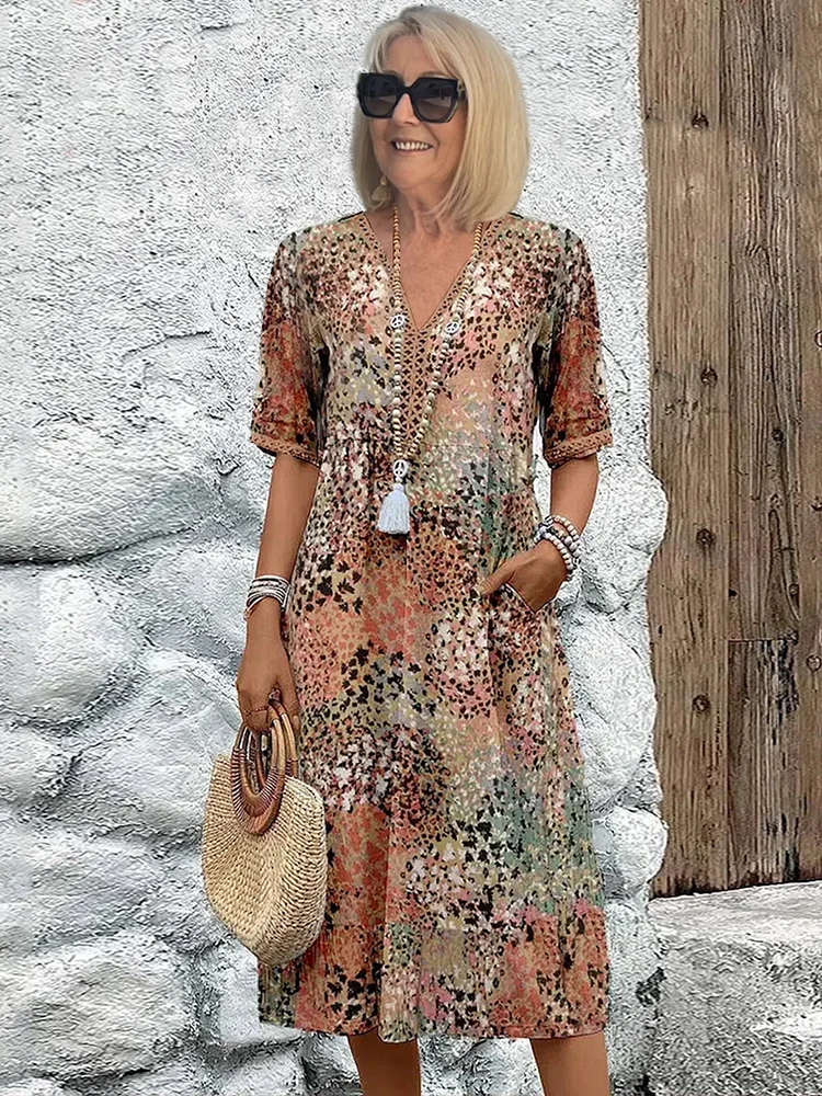 Femme souriante en robe d'été fleurie, lunettes de soleil, tenant un sac en paille, posant devant un mur blanc texturé et une porte en bois.