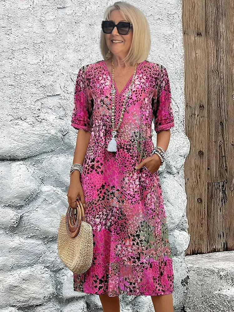 Femme souriante en robe rose à motifs floraux, portant des lunettes de soleil et un sac en paille, posant devant un mur texturé. Mode estivale élégante.