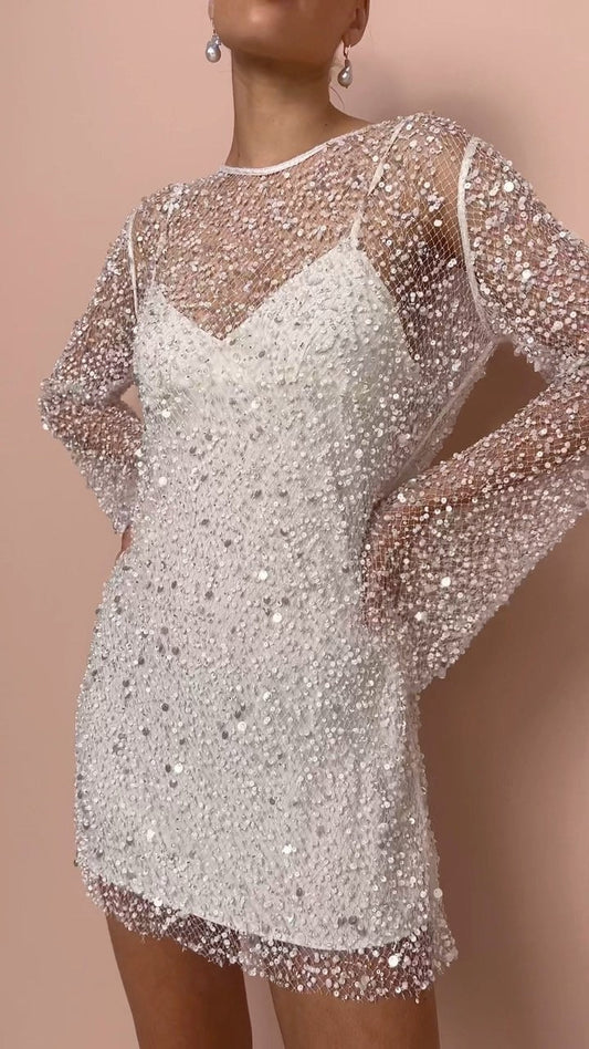 Robe blanche scintillante à manches longues en tulle transparent, ornée de paillettes, portée par une femme sur fond rose. Mode élégante et glamour.