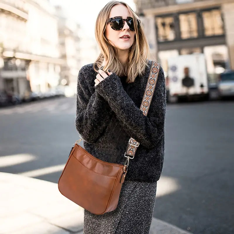 Femme portant un pull gris et un sac à bandoulière en cuir marron avec une sangle décorative, lunettes de soleil, rue urbaine en arrière-plan.