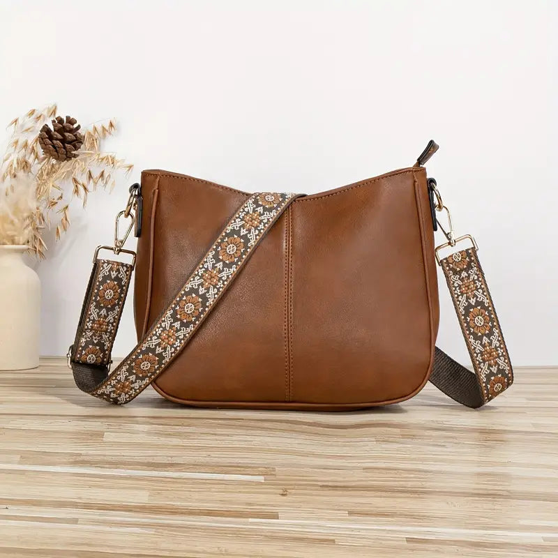 Sac à main en cuir marron avec bandoulière à motifs floraux, posé sur une table en bois clair, vase décoratif en arrière-plan. Accessoire mode tendance.