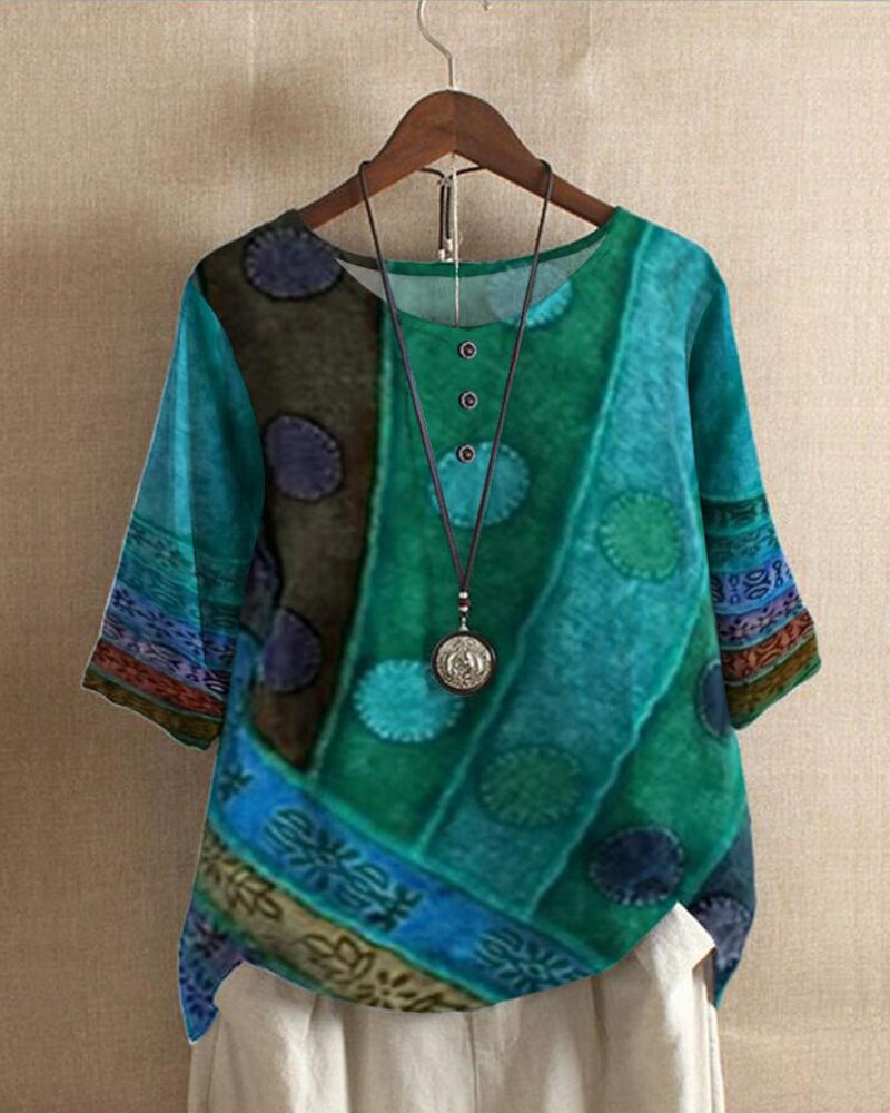 Blouse bohème verte à motifs ethniques, manches trois-quarts, col rond avec boutons, suspendue sur un cintre, collier pendentif argenté.