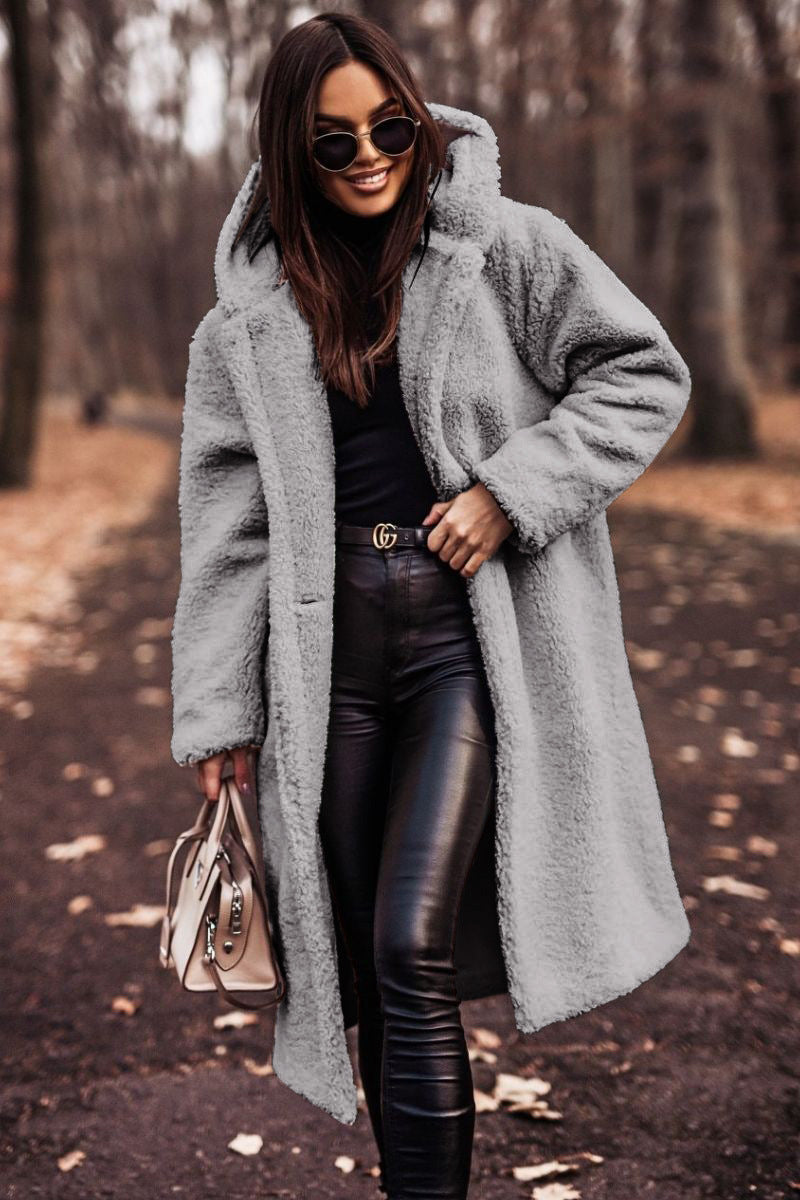 Femme portant un manteau en fausse fourrure gris, lunettes de soleil, pantalon en cuir noir, sac à main beige, marchant dans un parc automnal.