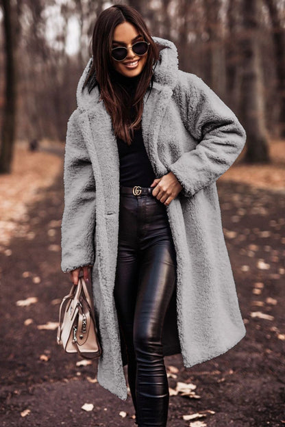 Femme portant un manteau en fausse fourrure gris, lunettes de soleil, pantalon en cuir noir, sac à main beige, marchant dans un parc automnal.