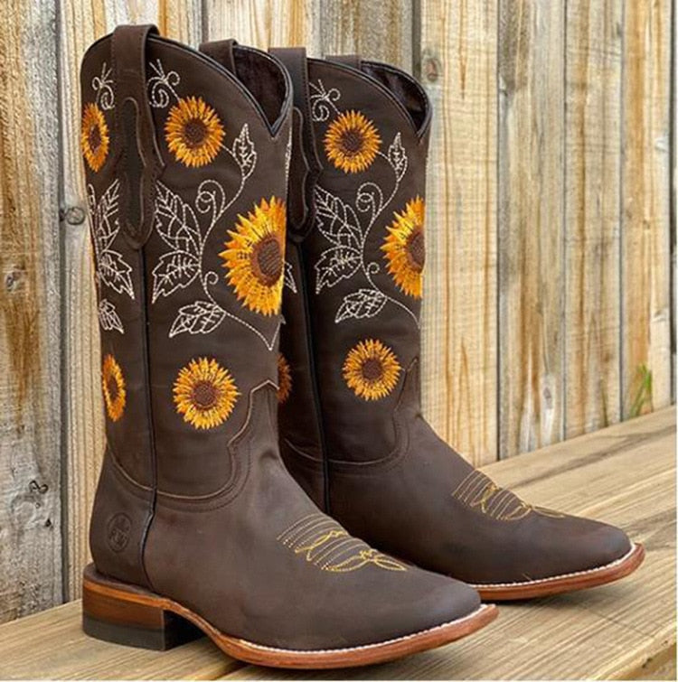 Brune cowboystøvler med solsikke-broderi, læder, westernstil, stående på træterrasse foran hegn. Perfekt til countrymode og rodeo.