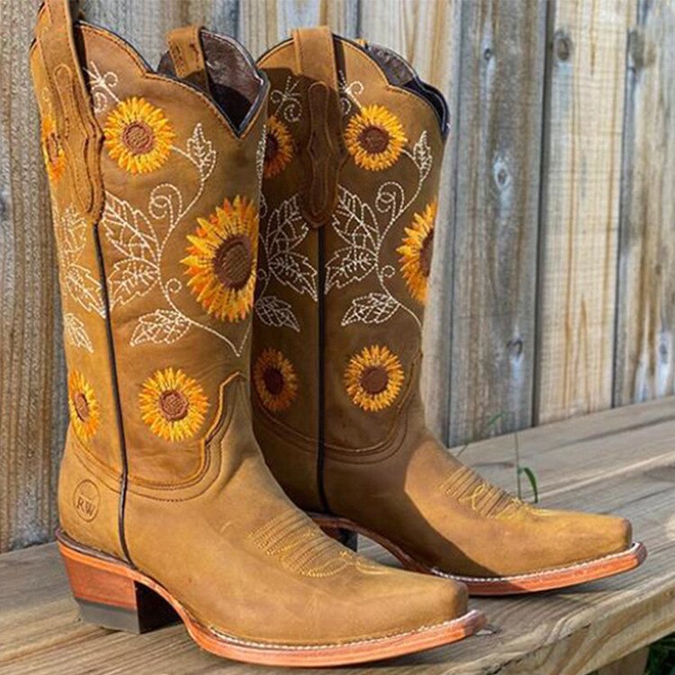 Brune cowboystøvler med solsikkemønster, broderede blade, træhegn i baggrunden. Perfekte til westernstil og landligt modeudtryk.