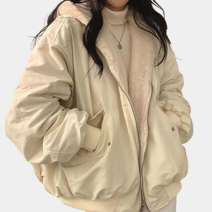 Dame i beige vinterjakke med hætte og pelsfor, iført en lys sweater og halskæde. Trendy overtøj, varm jakke, vintermode.