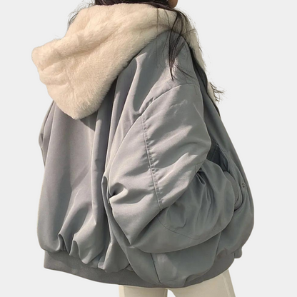 Kvinde iført grå, oversized bomberjakke med hvid pelskrave og hætte. Trendy vintermode, stilfuld overtøj, moderne design.