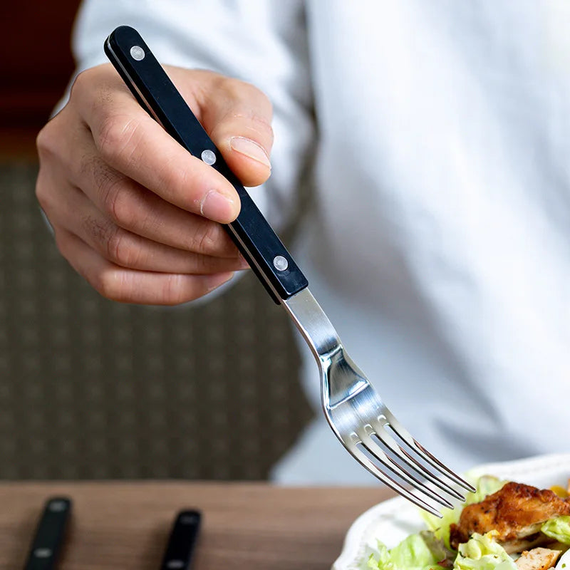 Hånd holder sort bestik gaffel over tallerken med salat og kød. Moderne køkkenredskab, rustfrit stål, ergonomisk design, spiseværktøj.