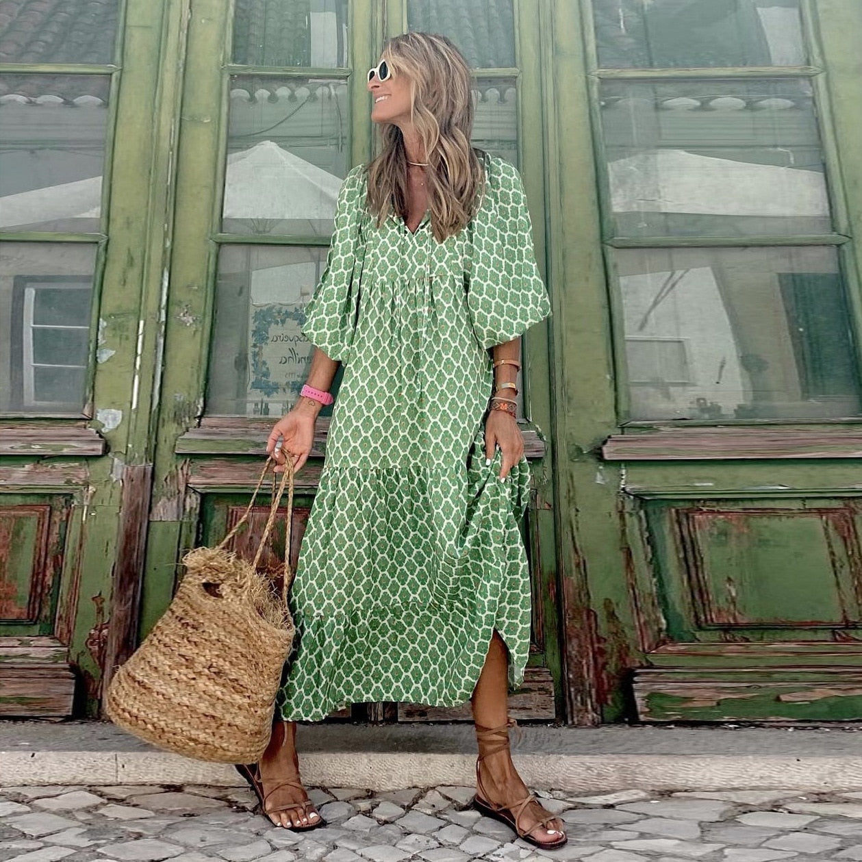 Frau in grünem Maxikleid mit geometrischem Muster, Strohtasche, Sandalen, vor grüner Holztür.