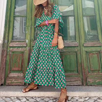 Frau in grünem Maxikleid mit rosa Muster, Strohhut und Sandalen vor grüner Holztür.