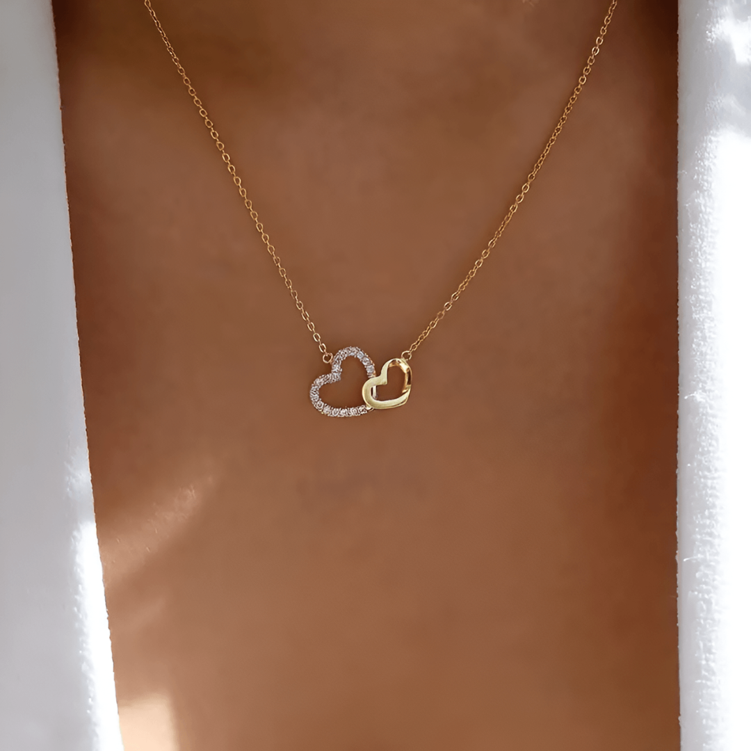 Goldene Halskette mit zwei Herzanhängern, einer mit Diamanten, auf Hauthintergrund.