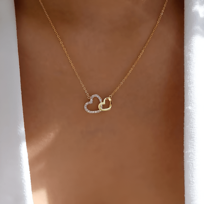 Goldene Halskette mit zwei Herzanhängern, einer mit Diamanten, auf Hauthintergrund.
