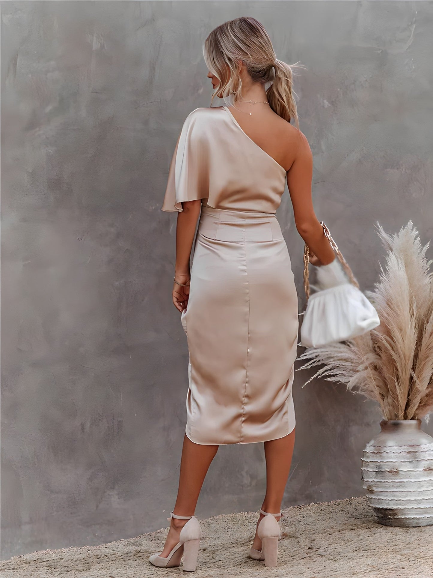 Femme en robe beige asymétrique élégante, tenant un sac blanc, posant près d'un vase décoratif avec des pampas, sur fond gris texturé.
