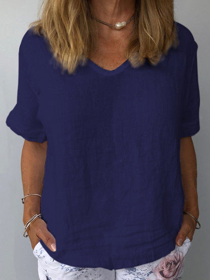 Femme portant un t-shirt bleu marine en lin, manches courtes, col en V, avec un collier perle et un pantalon blanc à motif floral. Mode décontractée élégante.