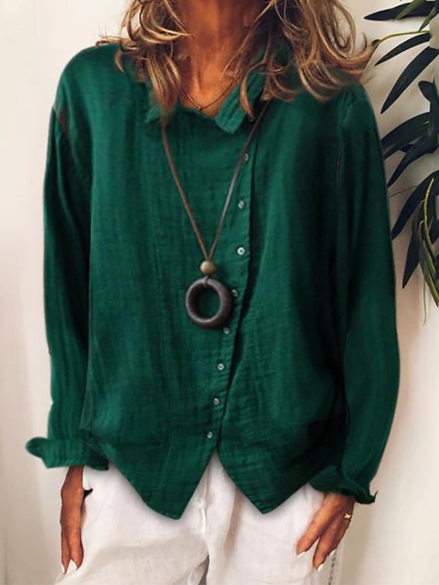 Femme portant une chemise verte ample avec un collier pendentif circulaire, posant devant un mur blanc avec des plantes. Mode décontractée élégante.