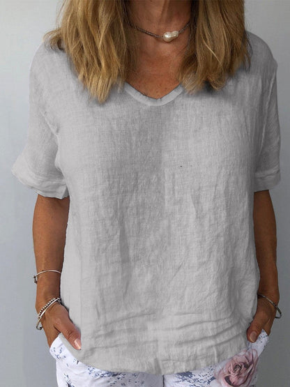 Femme portant un t-shirt en lin gris à col en V, manches courtes, avec un collier perle et un pantalon blanc à motif floral. Mode décontractée élégante.