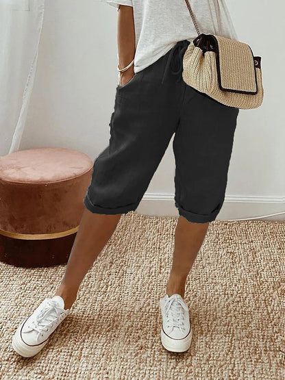 Femme portant un short noir décontracté, t-shirt blanc, baskets blanches et sac à main en paille, debout sur un tapis beige. Mode estivale chic.