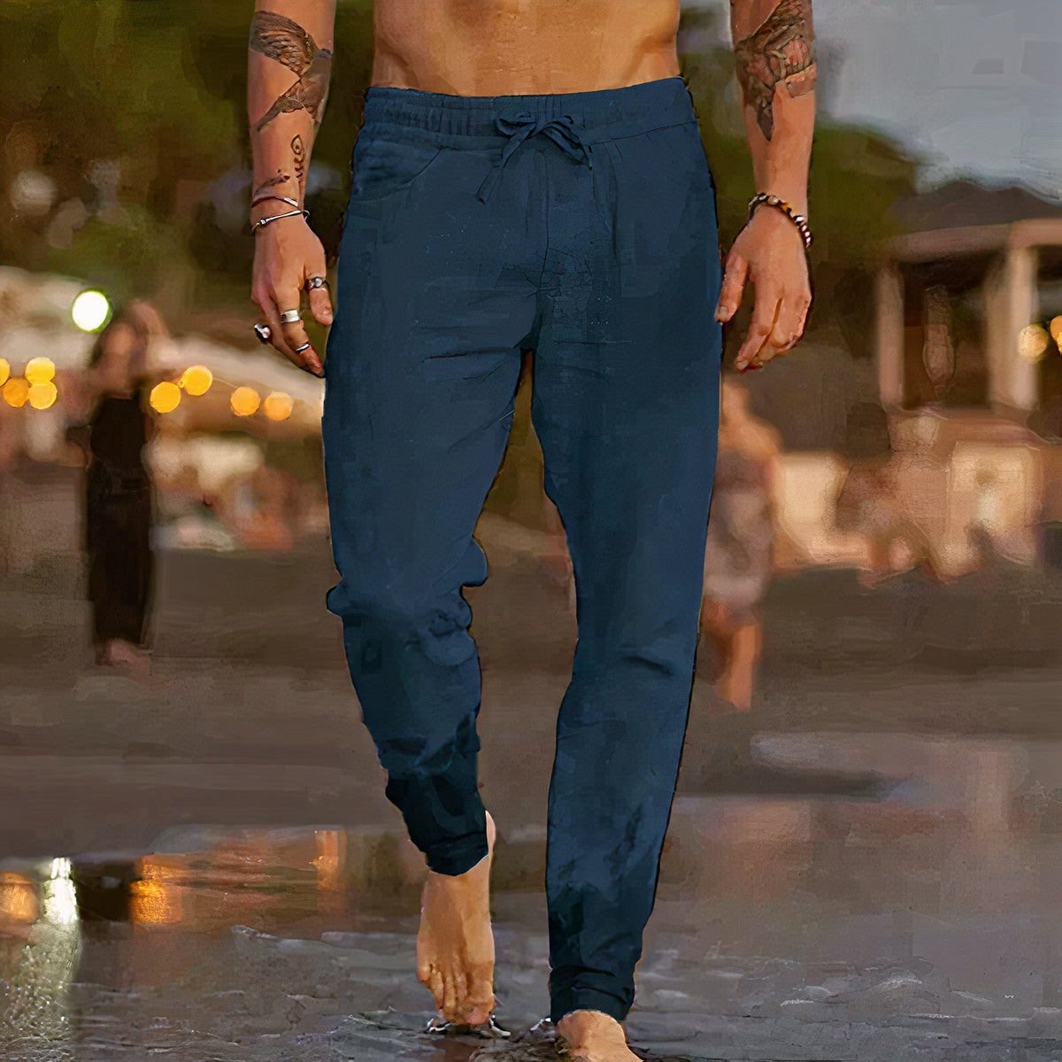 Homme torse nu portant un pantalon de jogging bleu foncé, marchant sur une plage au coucher du soleil, ambiance estivale et décontractée.